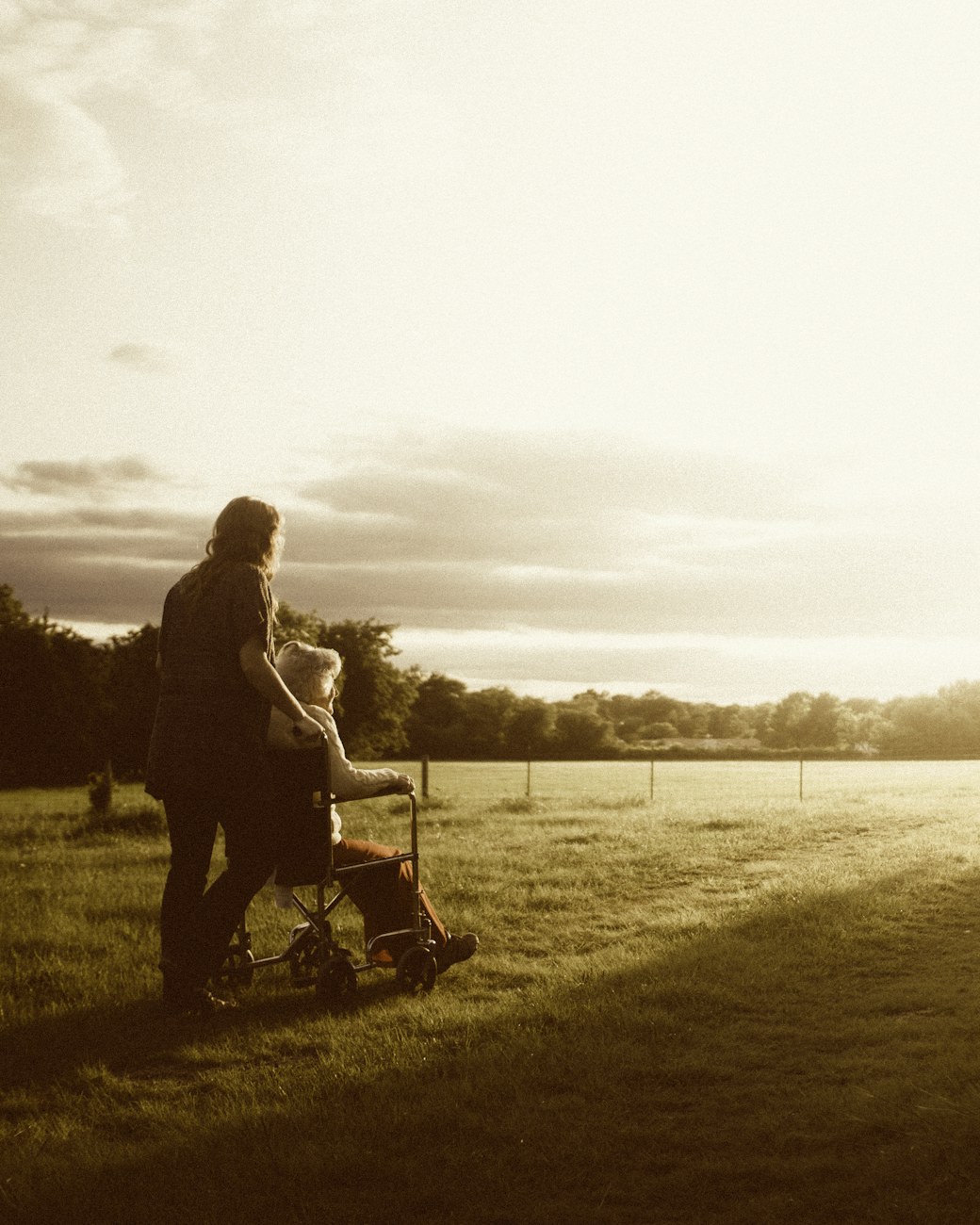 A caregiver standing beside an elderly woman, looking out over a sunlit field